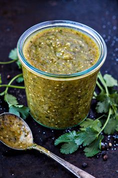 a glass jar filled with green salsa next to a spoon and cilantro sprigs