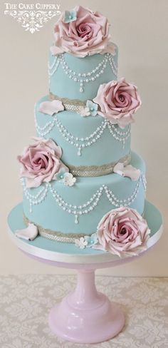 a three tiered blue wedding cake with pink roses on the top and pearls around the edges