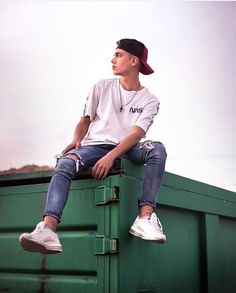 a man sitting on top of a green dumpster next to a red ball cap