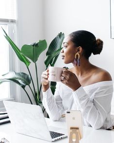 a woman sitting at a desk with a laptop and cup in her hand while holding a mug