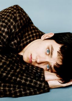 a man with long hair and blue eyes is laying on the floor wearing a brown coat