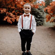 a young boy wearing suspenders and a bow tie standing in front of some trees