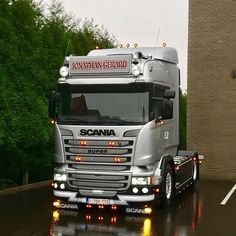 a silver scana truck driving down a rain soaked street in front of a brick building