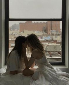 two young women sitting on a bed in front of a window, one kissing the other