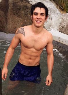 a shirtless man standing in the water next to a rock waterfall and smiling at the camera