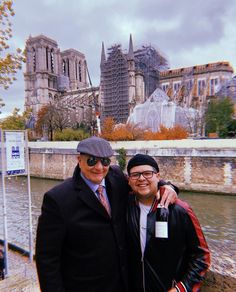 two men standing next to each other in front of a river with buildings behind them