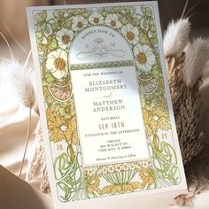 an ornate wedding card with flowers and leaves on it sitting next to some dried feathers