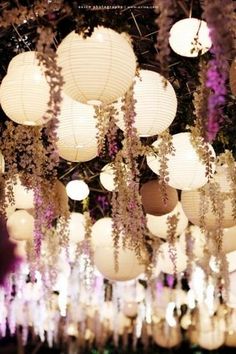 many white lanterns hanging from the ceiling with purple flowers and greenery all around them