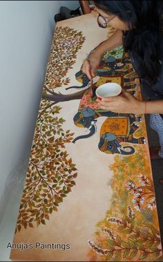 a woman is painting an elephant design on a large table cloth with white bowls in front of her