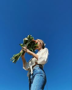 a woman is holding flowers in her hands and looking up into the sky with blue skies behind her