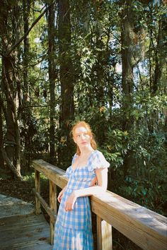 a woman standing on a wooden bridge in front of some trees and bushes, wearing a blue gingham dress