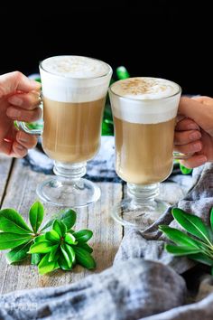 two people holding glasses filled with drinks on top of a wooden table next to green plants