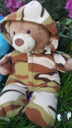 a teddy bear wearing a camouflage outfit laying in the grass with purple flowers behind it