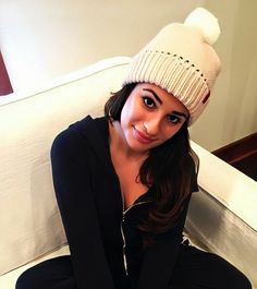 a woman sitting on a bed wearing a white knitted hat and black sweater with pom - poms