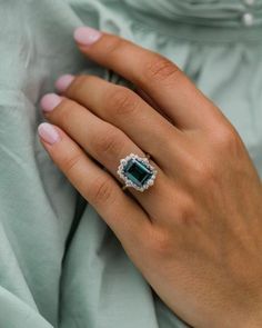 a woman's hand wearing a ring with an emerald stone in the center and diamond halo around it