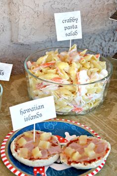 some food is sitting on a table with signs in front of it that read crabby pasta salad and hawaiian islands