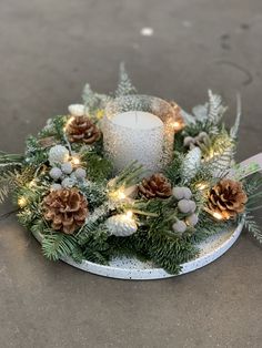 a white candle surrounded by pine cones and greenery with lights on the top is sitting on concrete