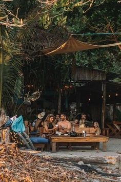 a group of people sitting around a wooden table on the beach in front of some trees