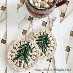 two crocheted earrings with green leaves on them sitting next to a mug filled with marshmallows