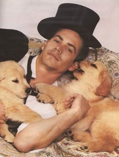 a man laying on top of a bed holding two puppies in his arms and wearing a black hat