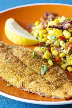 an orange plate topped with fried fish and corn salad next to a slice of lemon