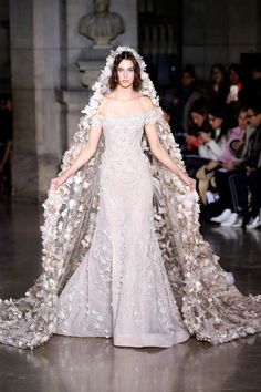 a model walks down the runway in a white gown with flowers on her cape and headdress