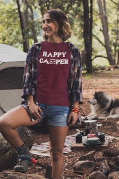 a woman standing in the woods next to a campfire with her dog looking on