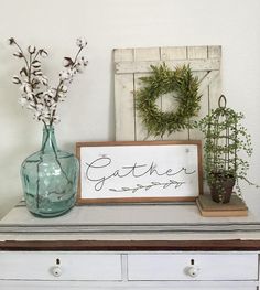 a white dresser topped with vases filled with flowers and greenery next to a sign that says gather