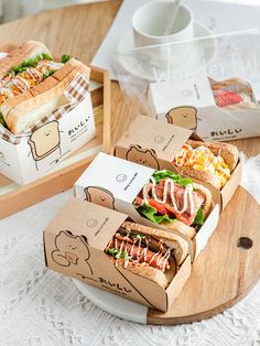 three sandwiches in boxes sitting on top of a wooden tray next to cups and saucers