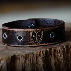 Made from premium grade 7 ounce leather. Comes engraved with the Crusader cross and shield shown centered between gunmetal eyelets. Strap is .75 inches wide. Hand dyed, distressed, oiled and finished in house. Made in America. Please note that adding personalization will replace the engraving shown. Instructions can be added in the note at checkout. Rustic Leather Concho Bracelets, Vintage Leather Concho Bracelets, Adjustable Leather Gothic Wristband, Vintage Leather Jewelry With Waxed Finish, Adjustable Gothic Leather Wristband, Vintage Leather Bracelet With Engraving, Vintage Stamped Leather Cuff Bracelet, Stamped Leather Vintage Cuff Bracelet, Vintage Engraved Leather Bracelets