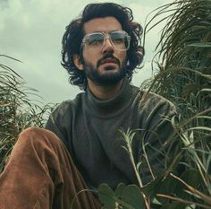 a man wearing glasses sitting in the middle of tall grass with palm trees behind him