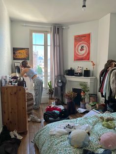 a woman standing next to a bed in a room filled with clothes and other items