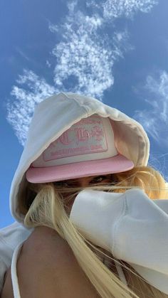 a blonde woman wearing a pink hat and white jacket with clouds in the sky behind her