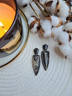 two earrings sitting next to a candle and some cotton on a white surface with a gold plate