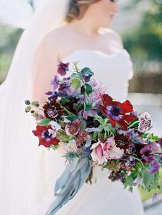 a bride holding a bouquet of purple and red flowers in her hand while wearing a veil