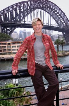 a man standing on top of a metal rail next to a body of water with a bridge in the background