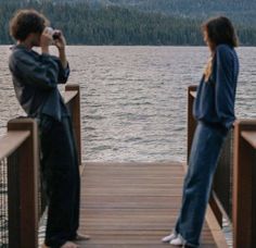 two people standing on a dock looking at the water and mountains in the distance, with one person taking a photo