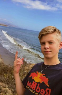 a young boy standing on top of a cliff near the ocean holding up his peace sign