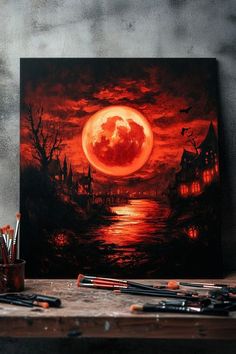 a painting on a table with paint brushes in front of it and an orange moon over the water