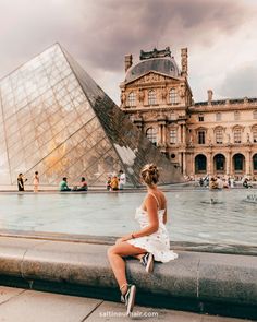 a woman sitting on the edge of a fountain in front of a building with a glass pyramid