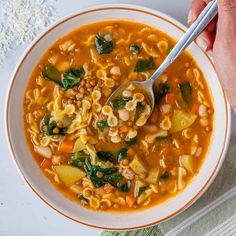 a person holding a spoon over a bowl of soup with noodles and spinach in it