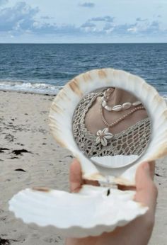 a person holding up a shell with jewelry in it on the beach next to the ocean