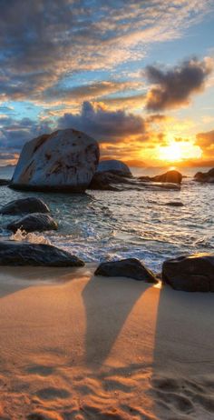 the sun is setting over some rocks in the water and there are footprints on the sand