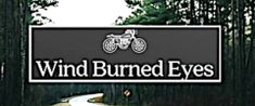 a sign that says wind burned eyes in front of some trees and dirt road with two motorcycles on it