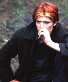 a man with red hair sitting on the ground in front of some bushes and trees