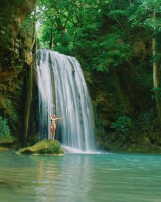 a woman standing on top of a rock in front of a waterfall with green foliage