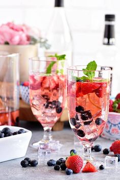 two glasses filled with fruit and ice on top of a table next to bowls of strawberries