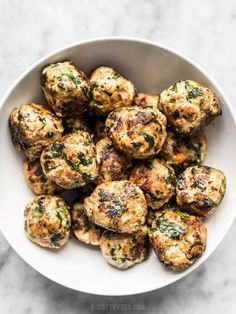 a white bowl filled with meatballs covered in cheese and spinach on top of a marble surface