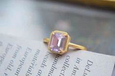 This ring features a central rectangular gemstone with a soft pink hue, cut in an emerald style with step facets that create a classic and elegant look. The stone is set in a gold band with four prongs securing it at each corner, and the setting is bordered by a decorative, textured frame that adds to the ring's vintage appeal. The overall design combines simplicity and sophistication, making it a charming and timeless piece. Details Isabella Rectangular Crystal Vintage Ring Adjustable opening Gold band Rare, antique design Reyis Jewelry Isabella Collection Gold Rectangular Crystal Ring, Elegant Gold Rectangular Crystal Ring, Elegant Ring With Faceted Rectangular Stone, Elegant Crystal Ring With Rectangular Stone, Formal Pink Rings With Rectangular Stone, Elegant Pink Rings With Rectangular Stone, Formal Pink Rectangular Ring, Gold Rectangular Amethyst Ring For Wedding, Gold Amethyst Rectangular Ring For Wedding