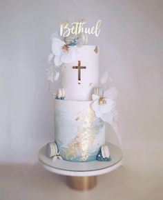 a three tiered cake with white and blue frosting, gold leaf decorations and a cross on top